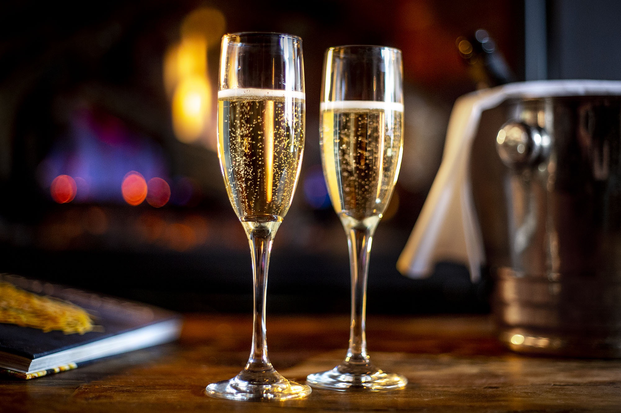 A photograph of two glasses of champaign in front of a fire.